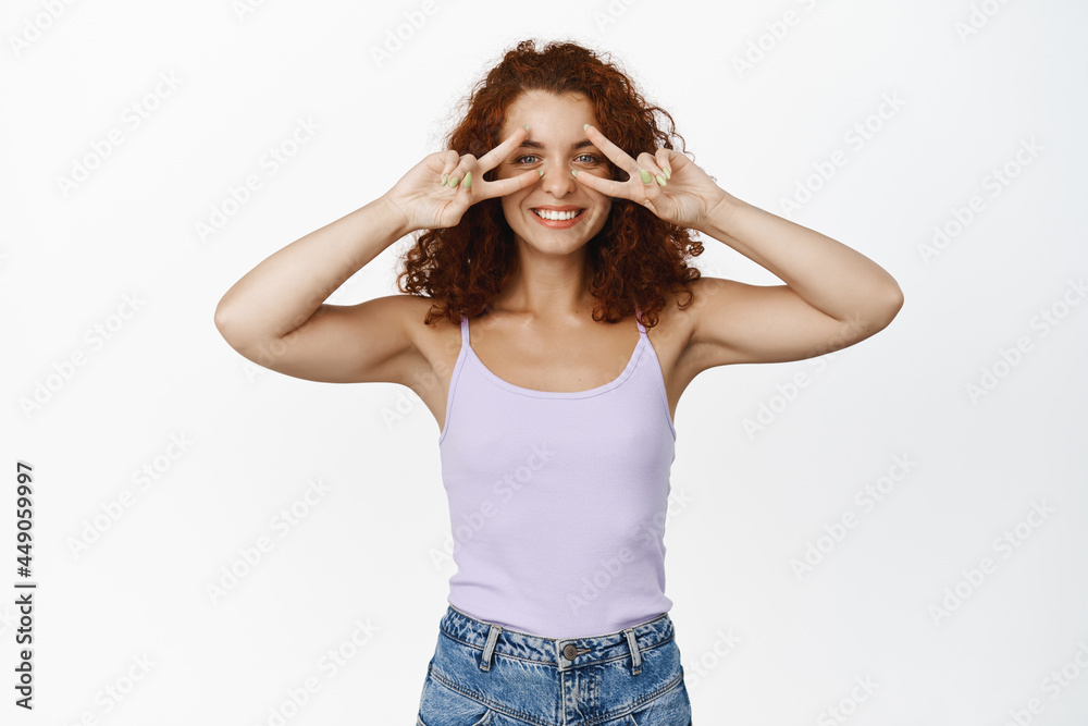 Positive redhead girl showing peace signs near eyes and smiling happy, cheerful mood, optimistic people and lifestyle concept, standing in summer clothes against white background Positive redhead girl showing peace signs near eyes and smiling happy, cheerful mood, optimistic people and lifestyle concept, standing in summer clothes against white background