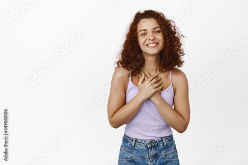 Thankful smiling redhead woman with curly hair, holding hands near chest and looking grateful, flattered with compliment, pleased of something good, white background