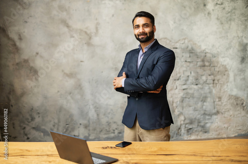 Canvas Print Indian businessman in formal suit stands in modern office near desk with a laptop and looking at camera