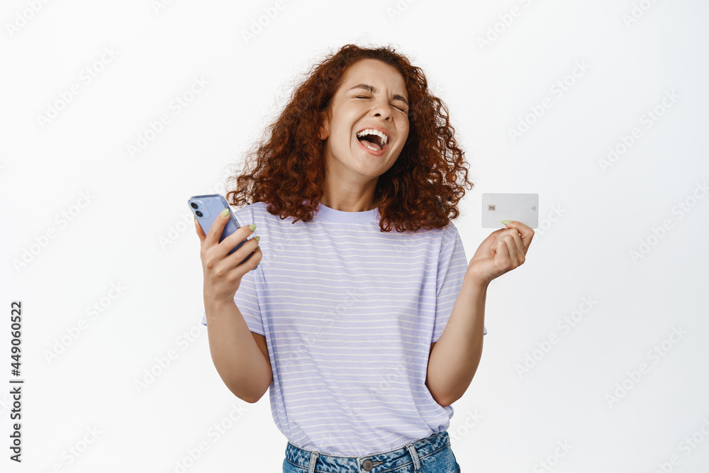 Excited curly girl laughing, holding smartphone with credit card, shopping online, making order in app, standing over white background happy Excited curly girl laughing, holding smartphone with credit card, shopping online, making order in app, standing over white background happy