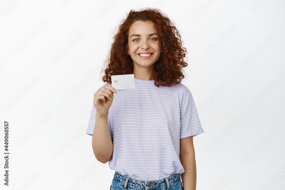 Stylish smiling redhead woman, showing her credit card and looking happy, become bank client, standing in casual t-shirt over white background Stylish smiling redhead woman, showing her credit card and looking happy, become bank client, standing in casual t-shirt over white background