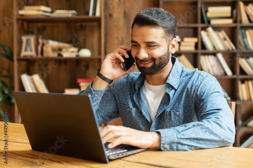 Mid adult indian man working in his home office, using smartphone and laptop for communication with customers or colleagues, smiling multiracial eastern guy talking phone watching on computer screen
