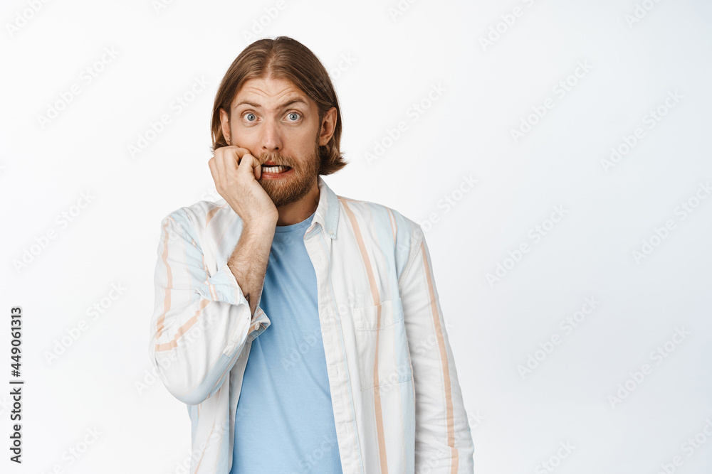 Portrait of anxious blond bearded guy biting fingers, gasping and looking startled at camera, terrified of something, watching smth scary, standing worried against white background