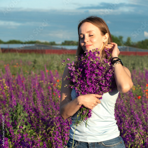 A fair girl with loose hair clasped to her a large bouquet of wild, bright lilac flowers, her eyes are covered, she straightens her hair with one hand, behind her is a flowering meadow.