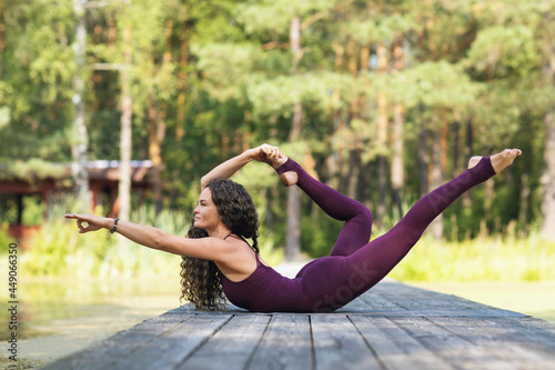 Beautiful sporty woman practicing yoga, on a wooden bridge in the park on a warm sunny morning, performs shalabhasana exercise, combining with dhanurasana