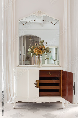 Vintage mirror and flower bouquet on wooden cupboard. Old restored furniture with opened door and drawers in classic bright interior with white wall and curtains