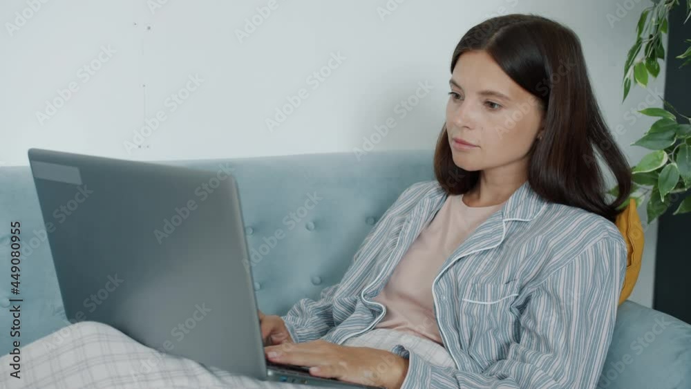 Pensive woman is working from home using laptop computer typing looking at documents sitting on comfy couch. Distant work and people concept.