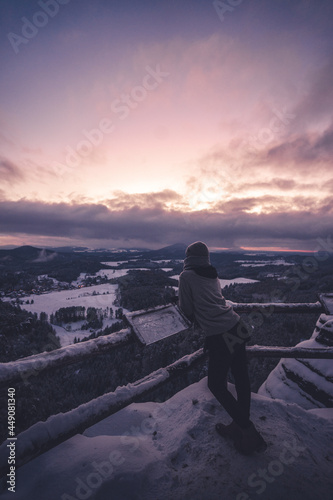 Woman leaning on the railing and watching how sun is going down. Dramatic wintry scene. Jetrichovice, Czech republic