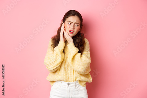 Young woman having toothache, touching swallen cheek and grimacing from pain, need to see dentist, stomatology appointment, standing against pink background photo