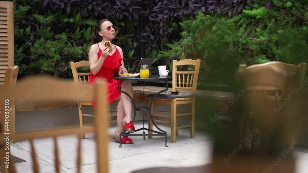 Wide shot of happy beautiful young Caucasian woman in red dress and sunglasses eating burger drinking cocktail sitting in cafe outdoors. Portrait of confident elegant freelancer with tablet on Wide shot of happy beautiful young Caucasian woman in red dress and sunglasses eating burger drinking cocktail sitting in cafe outdoors. Portrait of confident elegant freelancer with tablet on