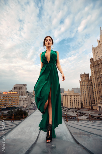 Attractive brunette girl in a long emerald dress posing on the catwalk on the roof against the background of the city, fashion concept, model in a luxurious dress against the background of urban photo