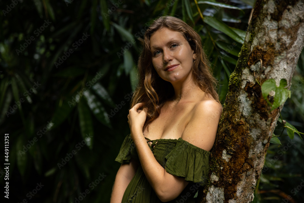 Portrait of Caucasian woman leaning against a tree in tropical rain forest. Beautiful smiling woman, wearing green dress. Nature concept. Travel to Asia. Bali, Indonesia Portrait of Caucasian woman leaning against a tree in tropical rain forest. Beautiful smiling woman, wearing green dress. Nature concept. Travel to Asia. Bali, Indonesia
