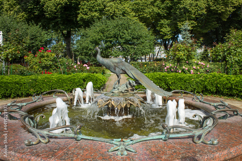 MOSCOW, RUSSIA-AUGUST, 4, 2021: a beautiful fountain with a bronze statue of a peacock and splashes of water among flower beds and lush foliage of green trees on a sunny summer day in a Kremlin park