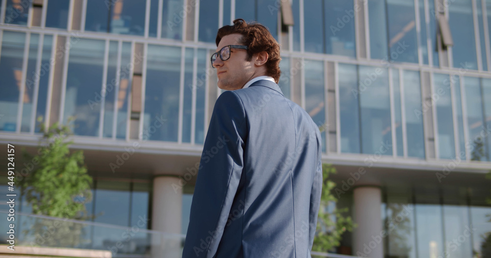 Back view of serious businessman in eye glasses looking up while standing against glass skyscraper