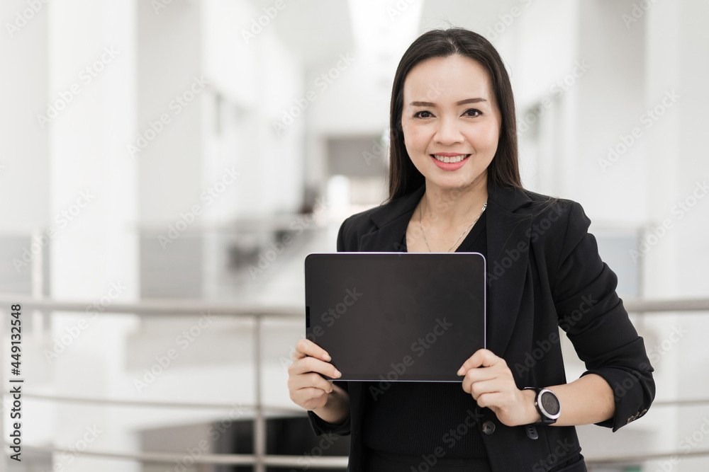 Portrait stock photo of a confident cheerful Asian businesswoman professional in black business suit using a digital tablet standing in the office building representing business oriented concept Portrait stock photo of a confident cheerful Asian businesswoman professional in black business suit using a digital tablet standing in the office building representing business oriented concept
