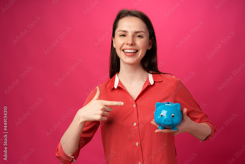 Happy smiling young woman holding blue piggy bank against pink background Happy smiling young woman holding blue piggy bank against pink background