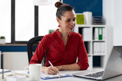 Smiling businesswoman reading data from laptop screen and taking notes on clipboard. Manager doing administrative wwork. Business leader doing financial expertise for client. photo
