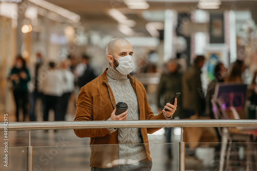 Man in a face mask is using a phone and holding a coffee in the shopping center.