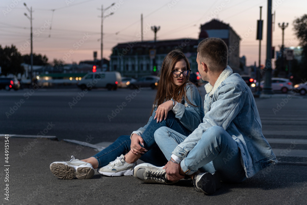 Modern young couple sit on the road on evening city background. Girl and a guy sort things out on the street.