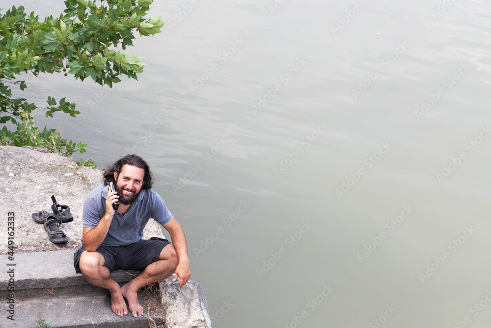 Smiling man with a beard barefoot on the steps against the background of trees and river speaks by phone,, color photo