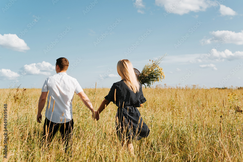 Happy young couple holding hands and walking in field, boyfriend and girlfriend in summer dress with bouquet of wild flowers, back view. Beautiful people in love enjoying time together. Love concept Happy young couple holding hands and walking in field, boyfriend and girlfriend in summer dress with bouquet of wild flowers, back view. Beautiful people in love enjoying time together. Love concept