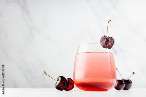 Cold fresh cherry beverage with sweet liqueur in misted glass decorated cherry on soft light white wood table, marble wall in modern interior.