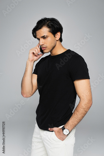 young man talking on cellphone and posing with hand in pocket isolated on grey