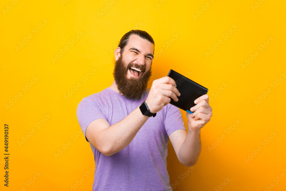 Excited young bearded man standing isolated over yellow wall background playing games his new tablet