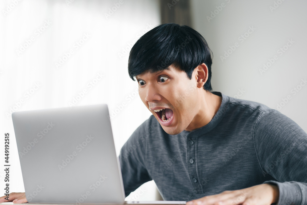 Shocked surprised Asian man looking at laptop computer screen sitting in the living room. Shocked surprised Asian man looking at laptop computer screen sitting in the living room.