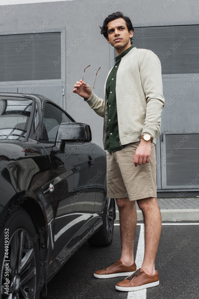 full length of young man holding stylish sunglasses and standing near modern car outside