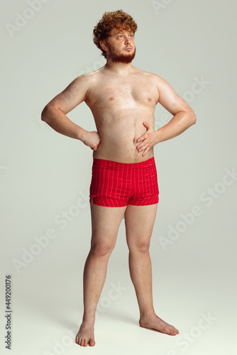 Cute red-headed man in red swimming shorts posing isolated on gray studio background. Concept of sport, humor and body positive.