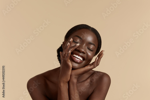 Portrait close up of beautiful african girl with closed eyes. Smiling young woman touch her clean face. Concept of face skin care. Isolated on beige background. Studio shoot