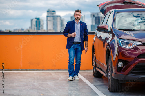Stylish man in a jacket and blue jeans posing in an open parking lot. Car parking in the background. Business center