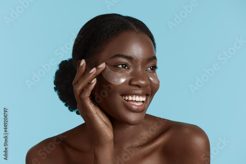 Close up portrait of beautiful african girl with under eye patch on face. Smiling young woman with perfect skin. Concept of face skin care. Isolated on blue background. Studio shoot