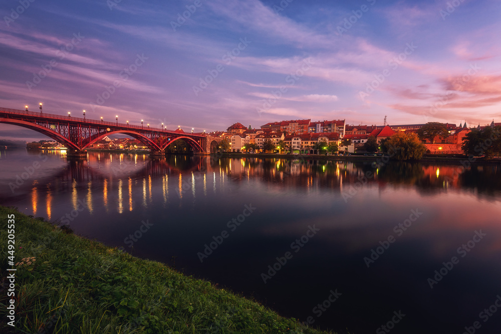 Obraz premium Amazing view of Maribor Old city, Main bridge (Stari most) on the banks of Drava river before sunrise, Slovenia. Scenic cityscape with dramatic cloudy sky and reflection, outdoor travel background