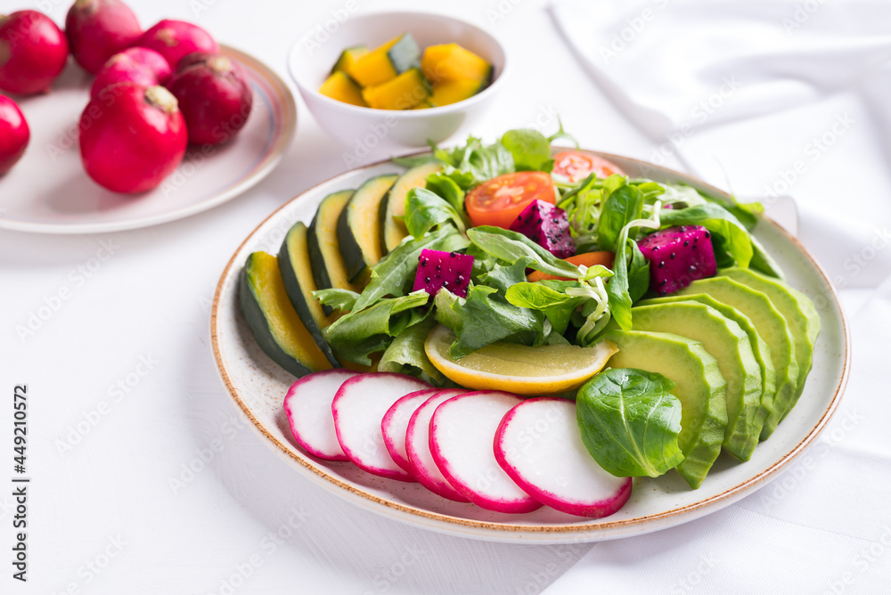 Healthy vegan salad fruits and vegetables with avocado, dragon fruit, radish, pumpkin, tomato, corn salad, lettuce, mizuna and lemon on white background