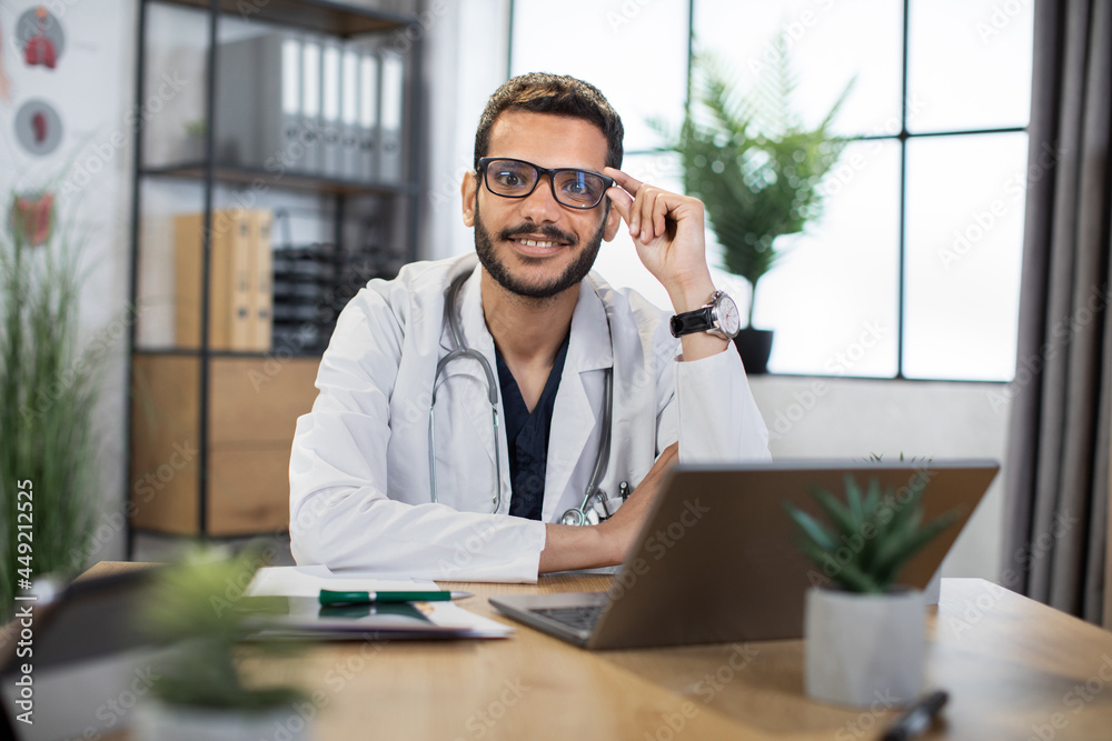 Close up of smiling handsome 30-aged male Malay doctor in eyeglasses, sitting at the table with laptop pc in his private office and looking at camera. Close up of smiling handsome 30-aged male Malay doctor in eyeglasses, sitting at the table with laptop pc in his private office and looking at camera.