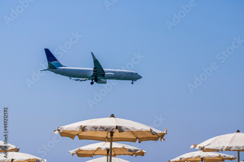passenger plane flying low above beach with blue sea with resting and swiming tourists and parasol umbrellas on the foreground