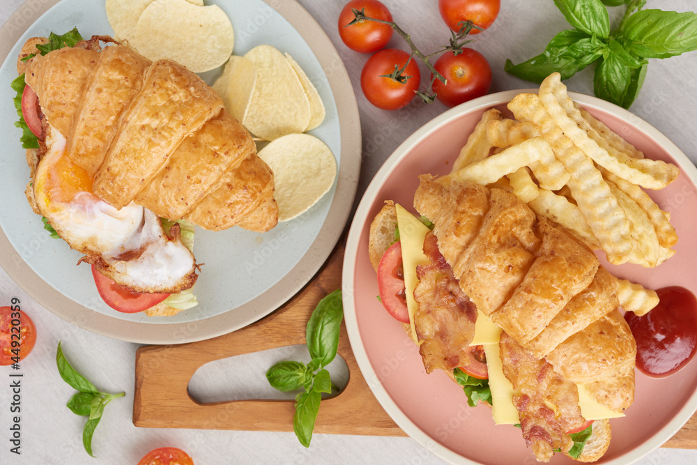 two croissant sandwiches on wooden table, top view, Sandwich with bacon, fried egg. Ham, cheese, bacon, fried egg, tomato, french fries and lettuce served in a plate.