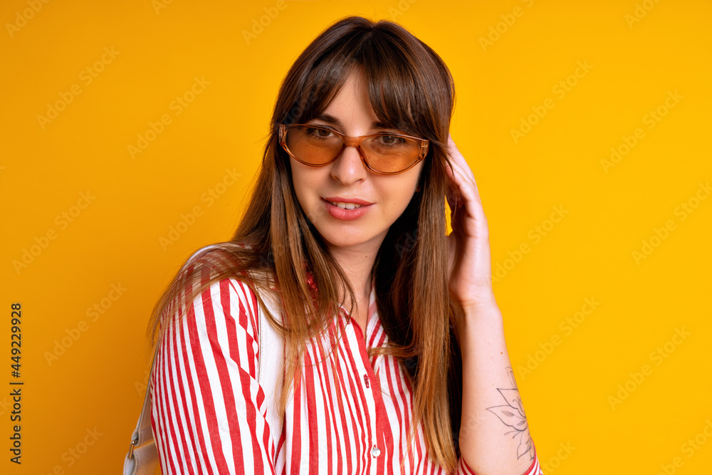 Fototapeta premium Cheerful pretty brunette woman wearing striped red dress, beige sunglasses and white bag, fashionable mood, studio yellow background.