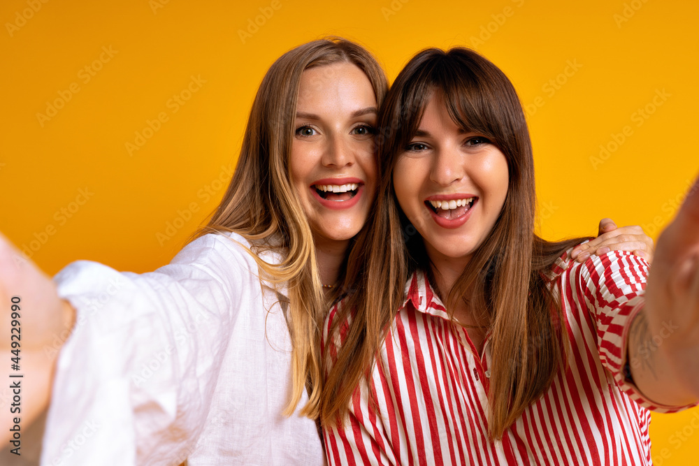 Close up studio portrait of happy best friends posing at studio yellow background, blonde and brunette having fun, cute couple.