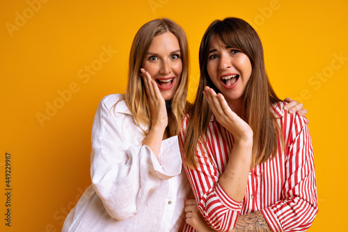 Close up studio portrait of happy best friends posing at studio yellow background, blonde and brunette having fun, cute couple.