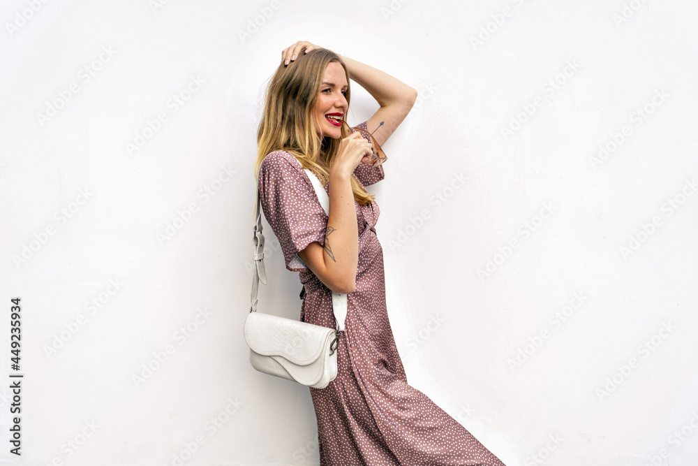 Fototapeta premium Indoor portrait of magnificent woman wearing elegant summer dress and accessorizes posing near white wall, happy emotions.
