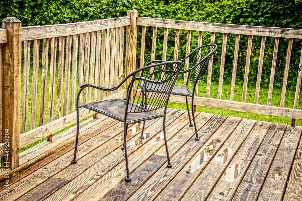 Two black metal lawn chairs sitting by rail on wooden deck after the rain with lush greenery in background.