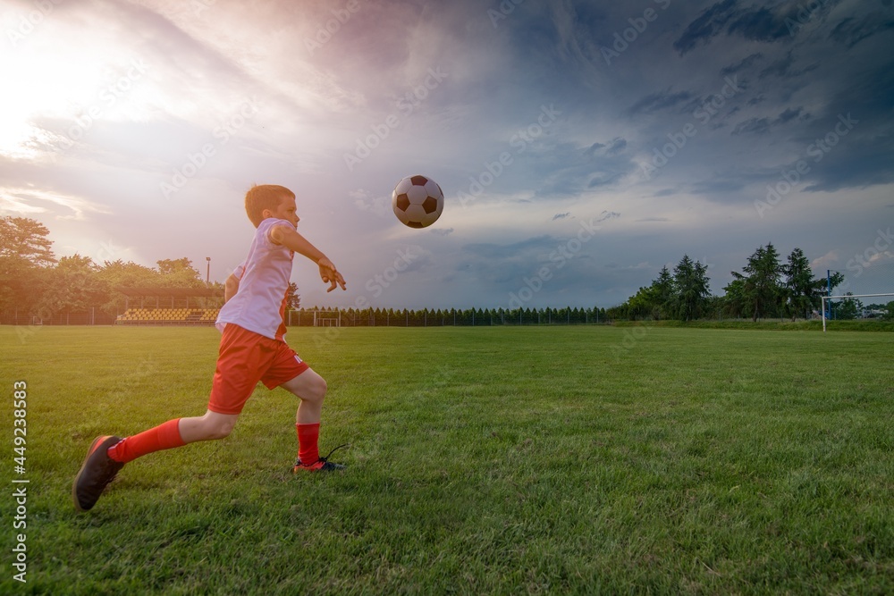 Boy playing with football ball on playing field. Child playing football. White - red football kit.