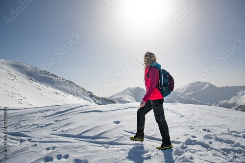 Female blonde tourist in a red jacket goes mountain trail in beautifull winter day.