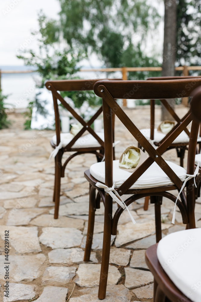 brown wooden chairs with crisscross back. White pillows on chairs and envelopes with white rose petals. against the backdrop of nature on a stone floor. holiday concept. brown wooden chairs with crisscross back. White pillows on chairs and envelopes with white rose petals. against the backdrop of nature on a stone floor. holiday concept.