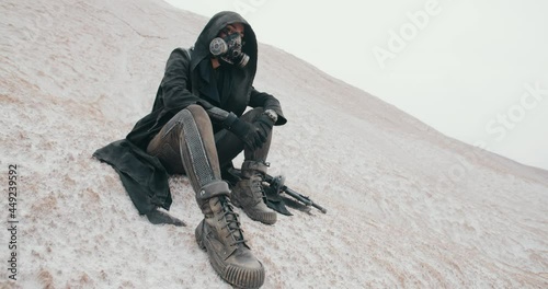 Wide angle post apocalyptic female in grunge hooded mantle and protective gas mask sitting on hill in dead valley desert. Steampunk cyberpunk soldier with gun. Future warrior after nuclear war photo