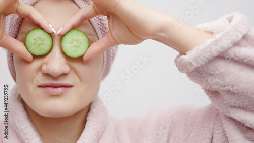 Attractive girl holding fresh cucumber slices and covering her eyes with the cucumber. The girl wearing a head towel and a pink bathrobe. Ready to take shower. Natural beauty and cosmetology concept.