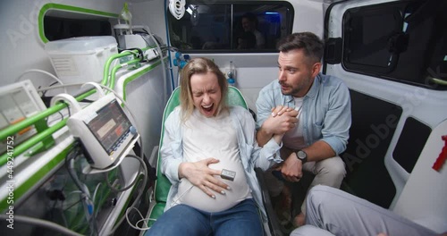 Caucasian young husband supporting pregnant wife in ambulance car during labor and taking selfie photos. Man breathing and holding woman hand when in childbirth. Making photo on way to hospital.